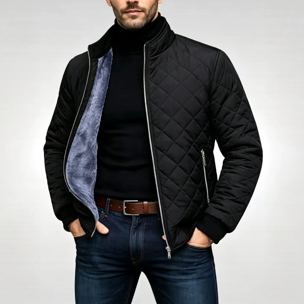 A man wearing a black quilted zip-up jacket with a gray fleece lining, styled with a black turtleneck, brown belt, and blue jeans against a light gray studio background.