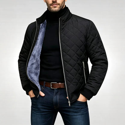 A man wearing a black quilted zip-up jacket with a gray fleece lining, styled with a black turtleneck, brown belt, and blue jeans against a light gray studio background.