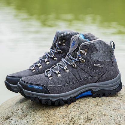 A pair of gray outdoor hiking boots with blue accents and silver metal eyelets, resting on a rock with a blurred body of water in the background.