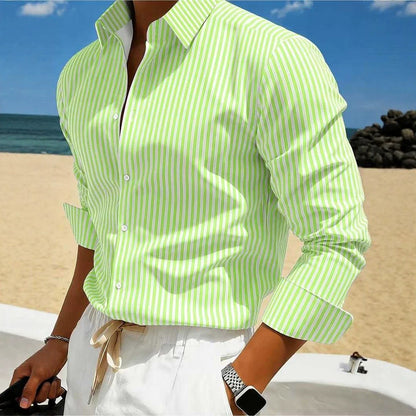 A man wearing a light green and white vertically striped long-sleeve button-up shirt, paired with white trousers, standing on a sandy beach with the ocean and blue sky in the background.