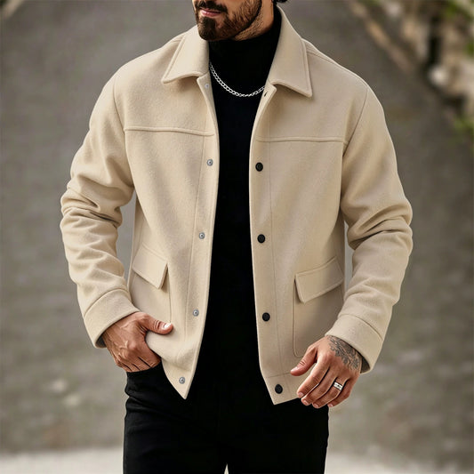 A man wearing a beige wool-blend button-up jacket over a black turtleneck, photographed outdoors against a blurred gray stone wall.