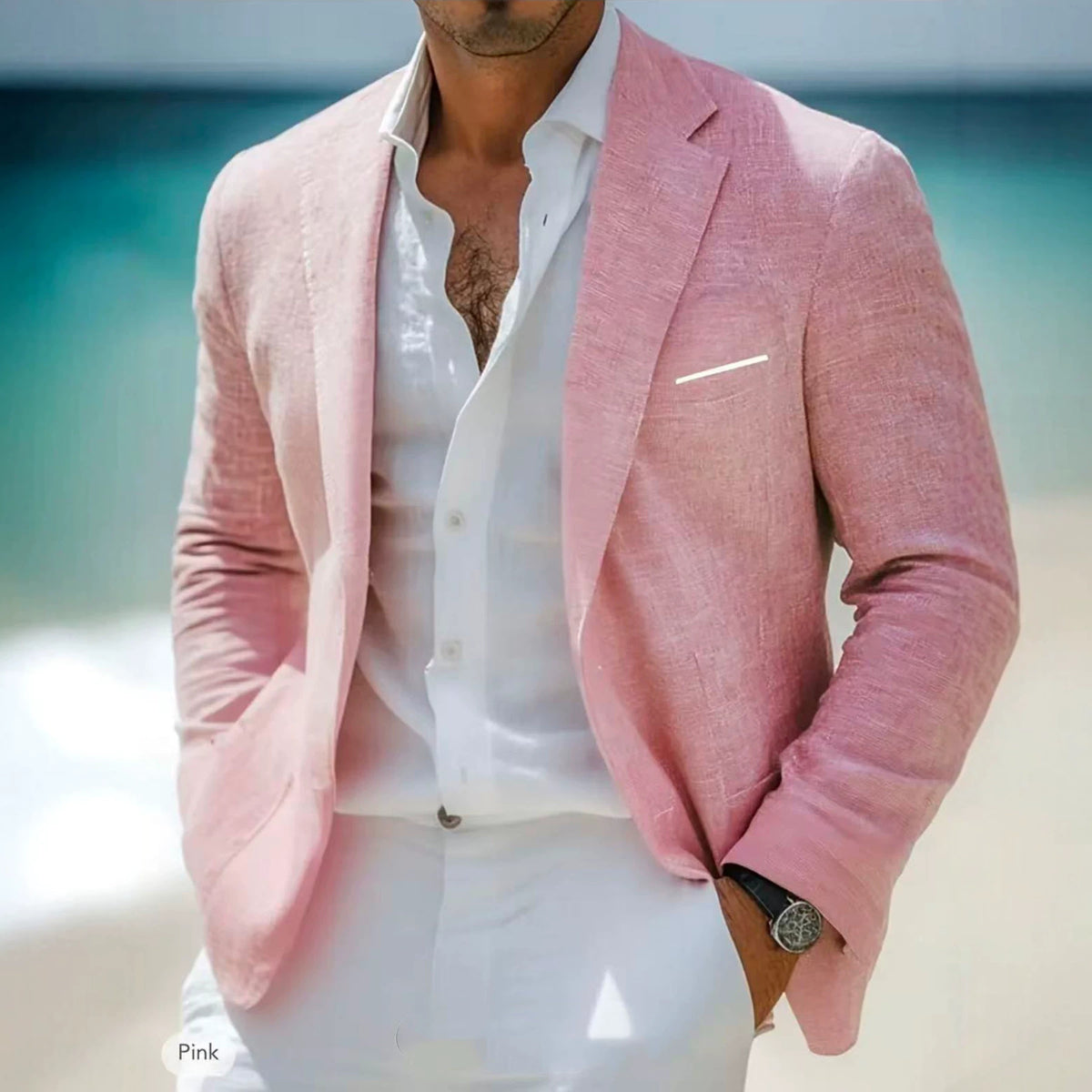 A man wearing a light pink textured blazer over a white dress shirt and white trousers, standing outdoors near a bright turquoise body of water.