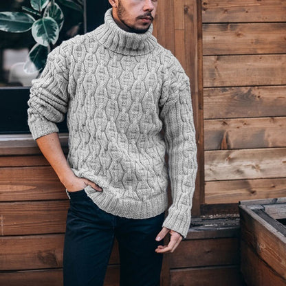 Man wearing a cable knit sweater standing against a wooden wall.