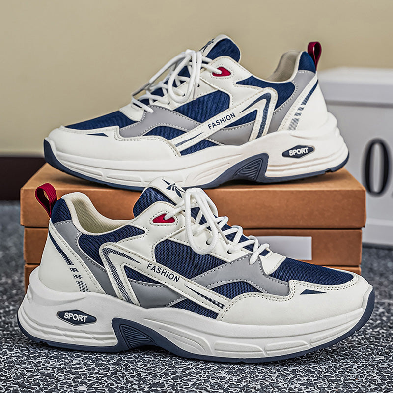 A pair of chunky low-top sneakers in navy blue, white, and gray, stacked on a brown shoebox. The shoes feature mesh panels, synthetic overlays, white laces, and small red accents on the tongue and heel pull tabs.
