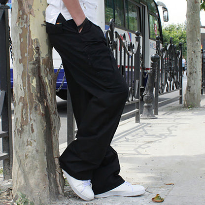 Person wearing black wide-leg pants and white sneakers leaning against a tree on a sidewalk.