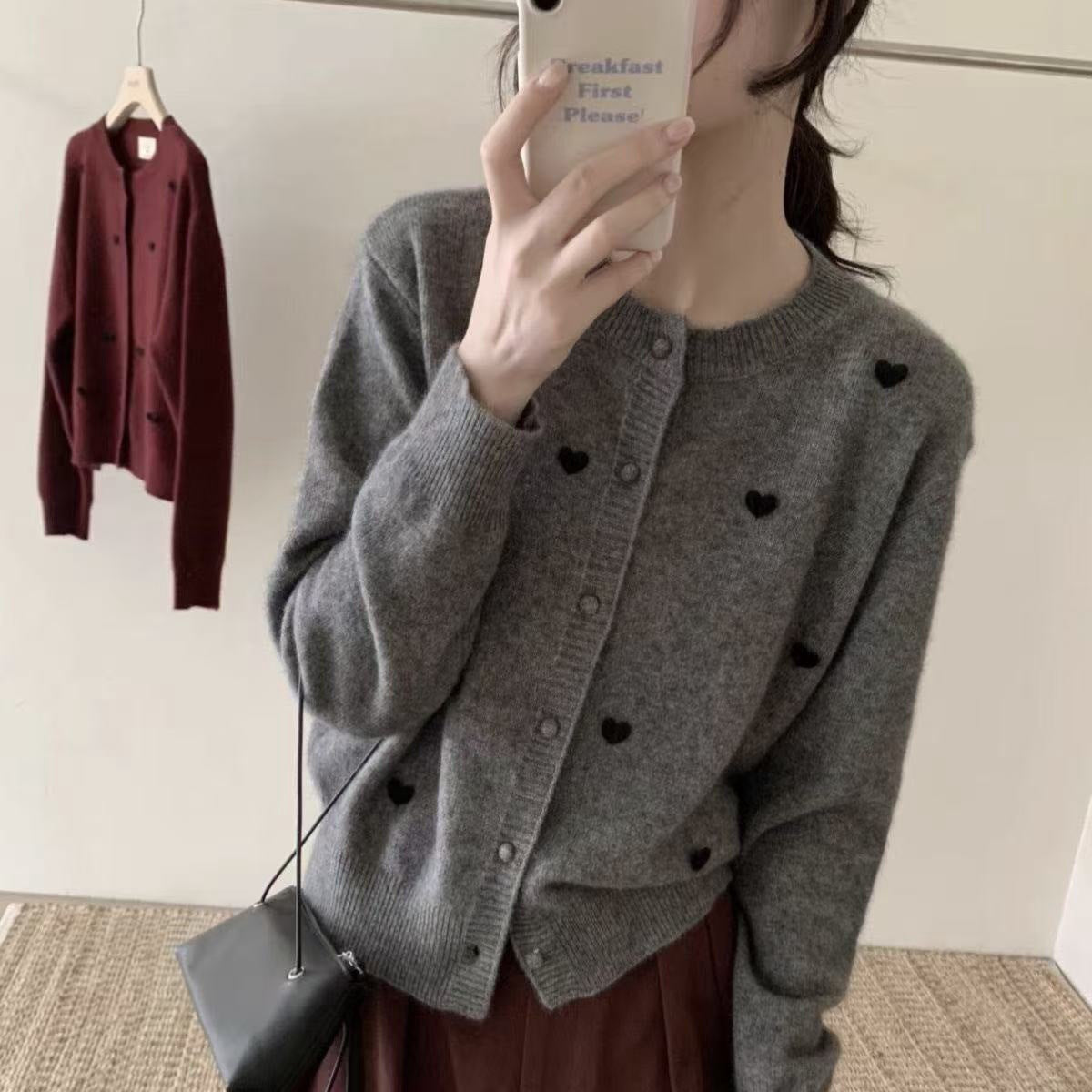 A person takes a mirror selfie wearing a gray knit cardigan with small black heart details, paired with a dark brown skirt. A matching dark brown cardigan is hanging on a hanger in the background.