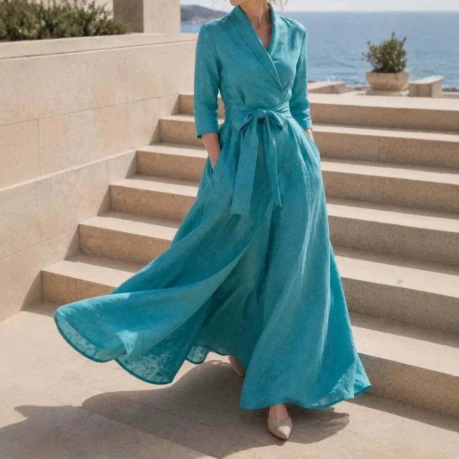A woman wearing a flowing turquoise maxi dress with three-quarter sleeves and a self-tie waist, walking down outdoor stone steps near the sea.