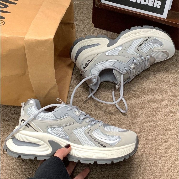 A pair of chunky low-top sneakers in light gray and white mesh with thick sculpted soles, shown on a carpeted floor with a brown paper bag and a shoe box in the background. A hand is holding one shoe.