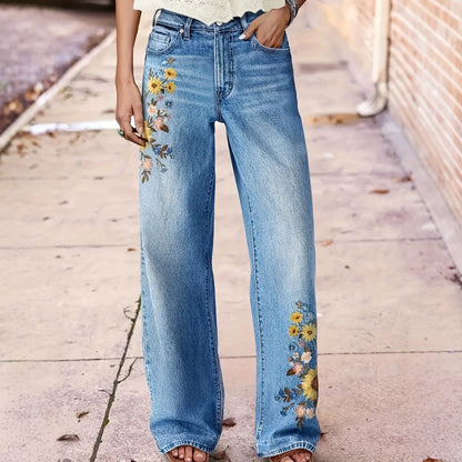 A person wearing light blue wide-leg denim jeans with floral embroidery on the left thigh and lower leg, standing on a sidewalk next to a brick wall.