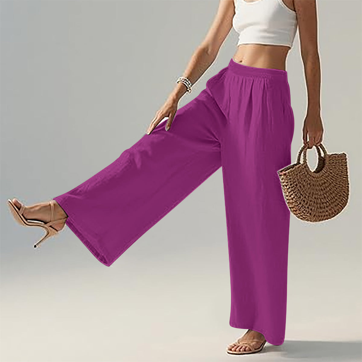 A model wearing bright magenta wide-leg pants, styled with a white crop top, beige high-heeled sandals, and a woven straw handbag against a light gray studio background.