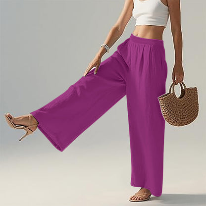 A model wearing bright magenta wide-leg pants, styled with a white crop top, beige high-heeled sandals, and a woven straw handbag against a light gray studio background.