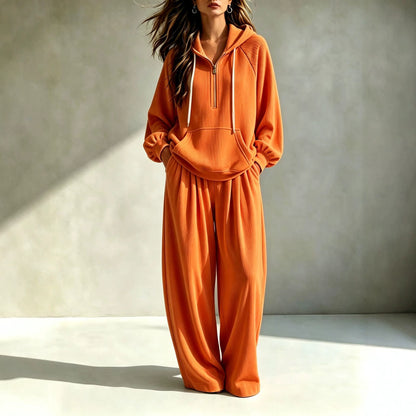 A woman models a matching orange loungewear set consisting of a half-zip hoodie and wide-leg sweatpants, standing against a textured gray wall with dramatic sunlight and shadows.