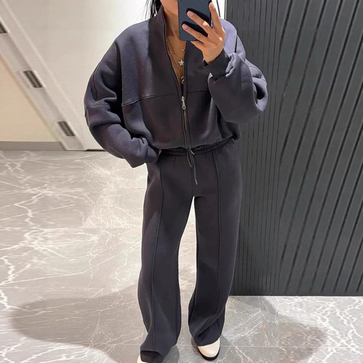 A person takes a mirror selfie wearing a dark gray matching zip-up jacket and wide-leg sweatpants set in a modern indoor space with a gray marble floor and dark paneled wall.