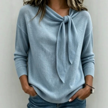 A woman wearing a light blue knit sweater with a large tie-neck detail, paired with blue jeans, shown against a plain light gray background.