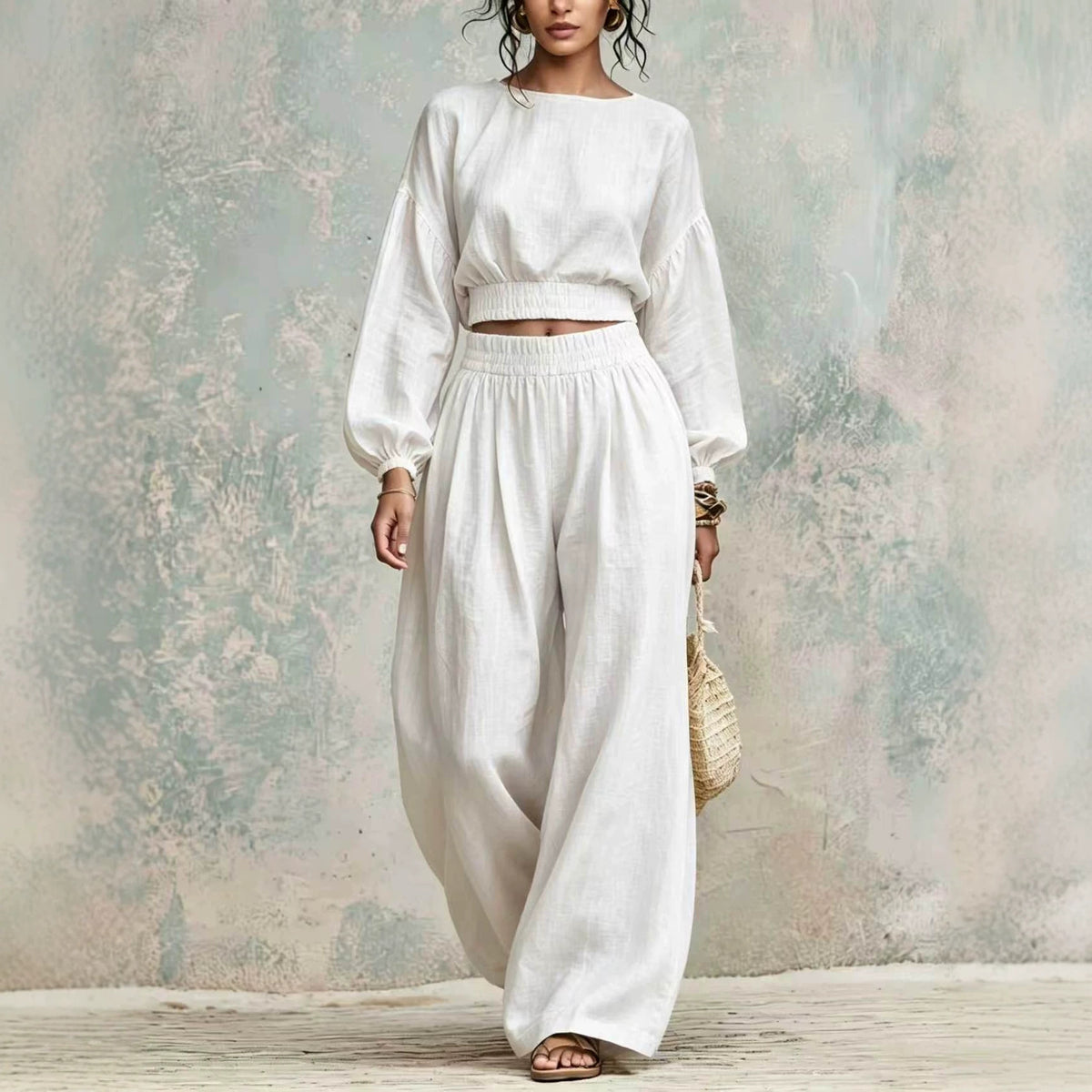 A woman models a white two-piece linen set consisting of a cropped long-sleeve top and matching wide-leg trousers, standing against a textured, rustic plaster wall.