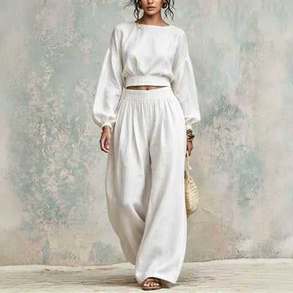 A woman models a white two-piece linen set consisting of a cropped long-sleeve top and matching wide-leg trousers, standing against a textured, rustic plaster wall.