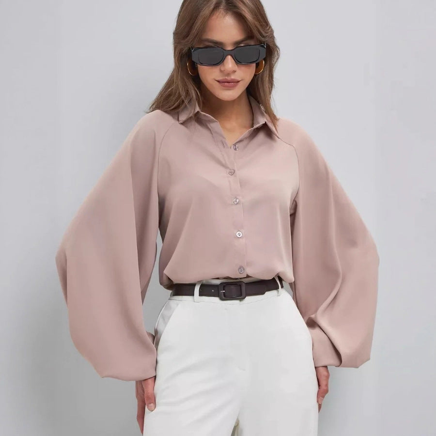 A woman wearing a dusty pink long-sleeve button-up blouse with dramatic balloon sleeves, paired with white trousers and a black belt, against a light gray studio background.