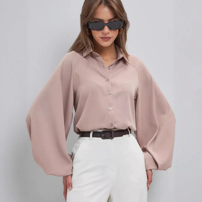 A woman wearing a dusty pink long-sleeve button-up blouse with dramatic balloon sleeves, paired with white trousers and a black belt, against a light gray studio background.