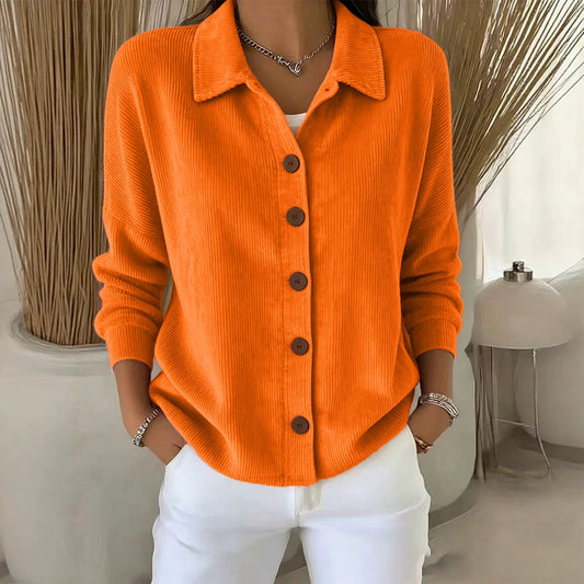 A woman wearing a bright orange ribbed knit button-up cardigan with a collar, styled with white pants and jewelry in a modern indoor setting.