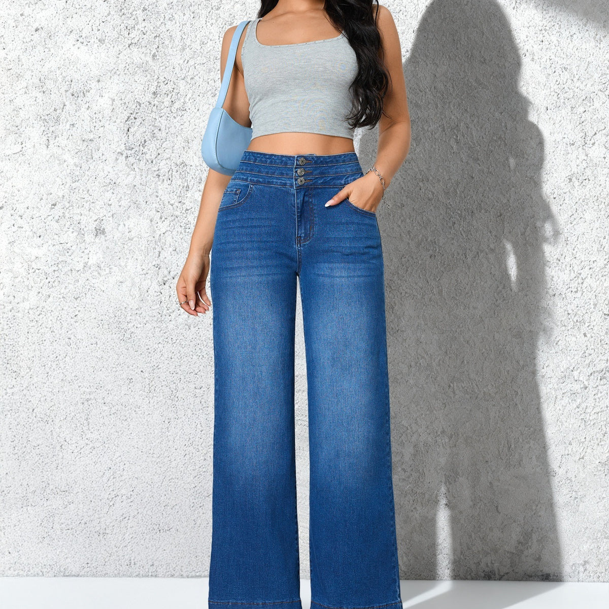 A model wearing blue wide-leg denim jeans with a light gray crop top and a light blue shoulder bag, standing against a textured gray wall with a strong shadow.